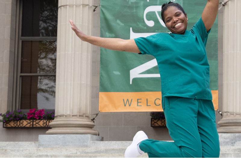 Smiling woman in green scrubs jumping with joy in front of a building with a